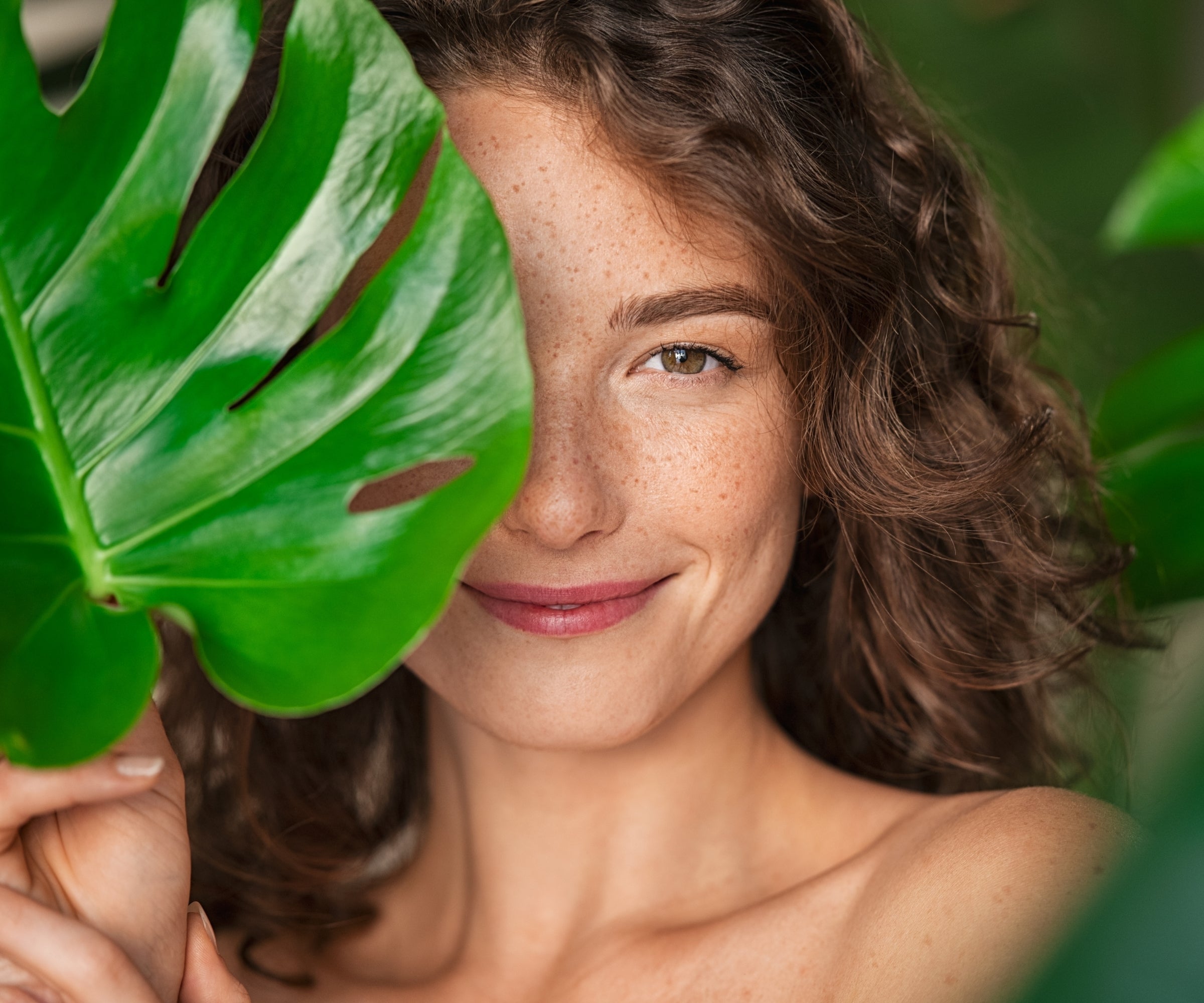 Mulher com pele natural e luminosa entre folhas verdes tropicais, representando beleza natural, skincare botânico e bem-estar inspirado na natureza em Estoril, Portugal.